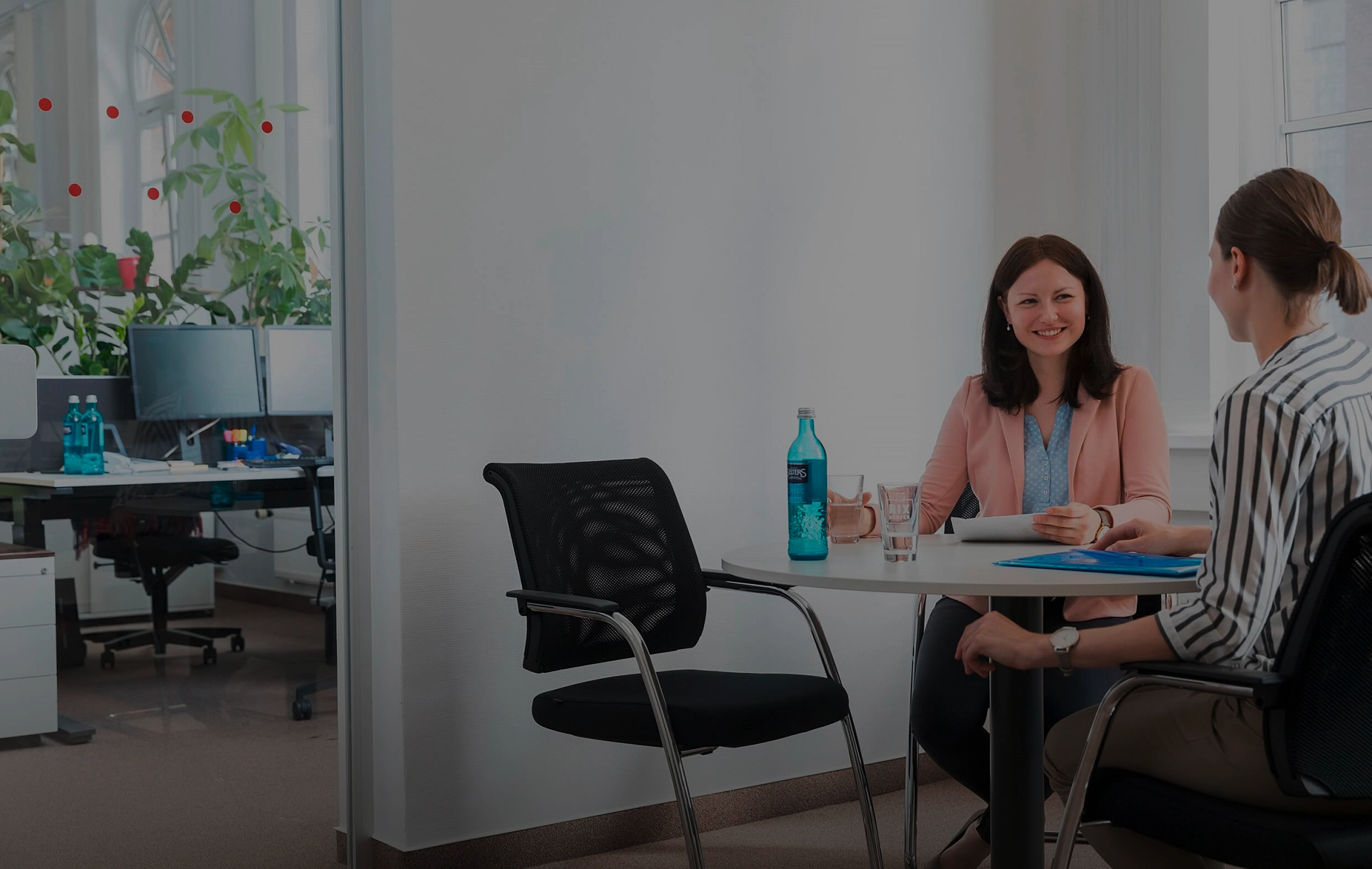 Zwei Frauen sitzen im Büro an einem Tisch und führen lächelnd ein Gespräch mit Unterlagen und Getränken.
