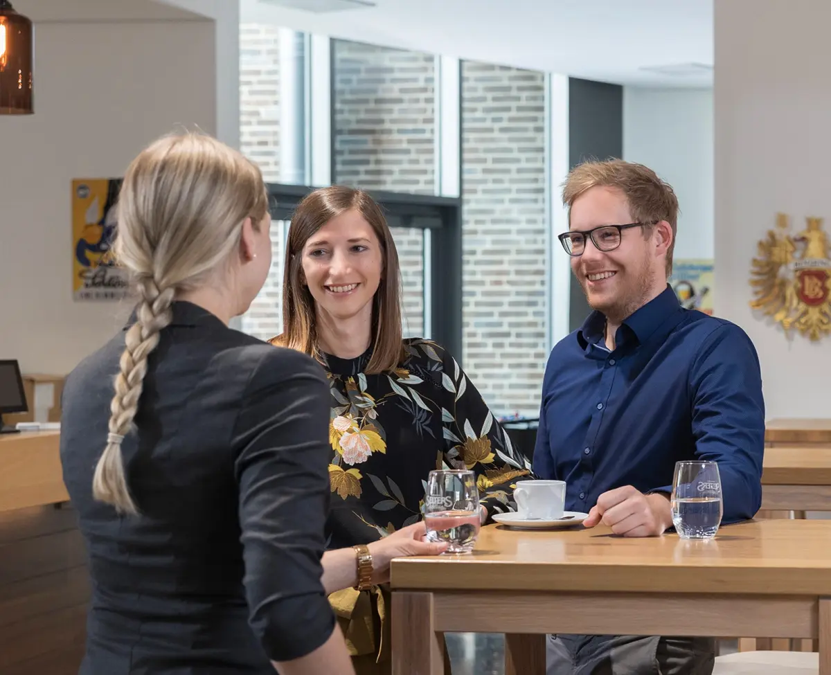Zwei Frauen und ein Mann sitzen lächelnd im Gespräch an einem Tisch mit Wassergläsern und einer Tasse.