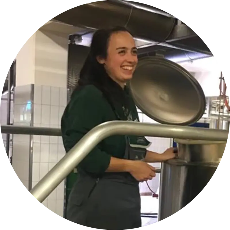 Eine junge Frau lächelt und steht neben einem geöffneten Edelstahlbehälter in einer Brauerei.