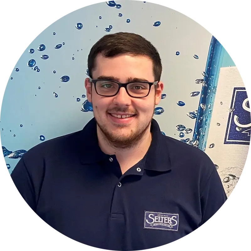 Ein junger Mann mit Brille und Polohemd steht vor einer Wand mit Wassertropfenmuster. Selters-Logo auf dem Hemd.