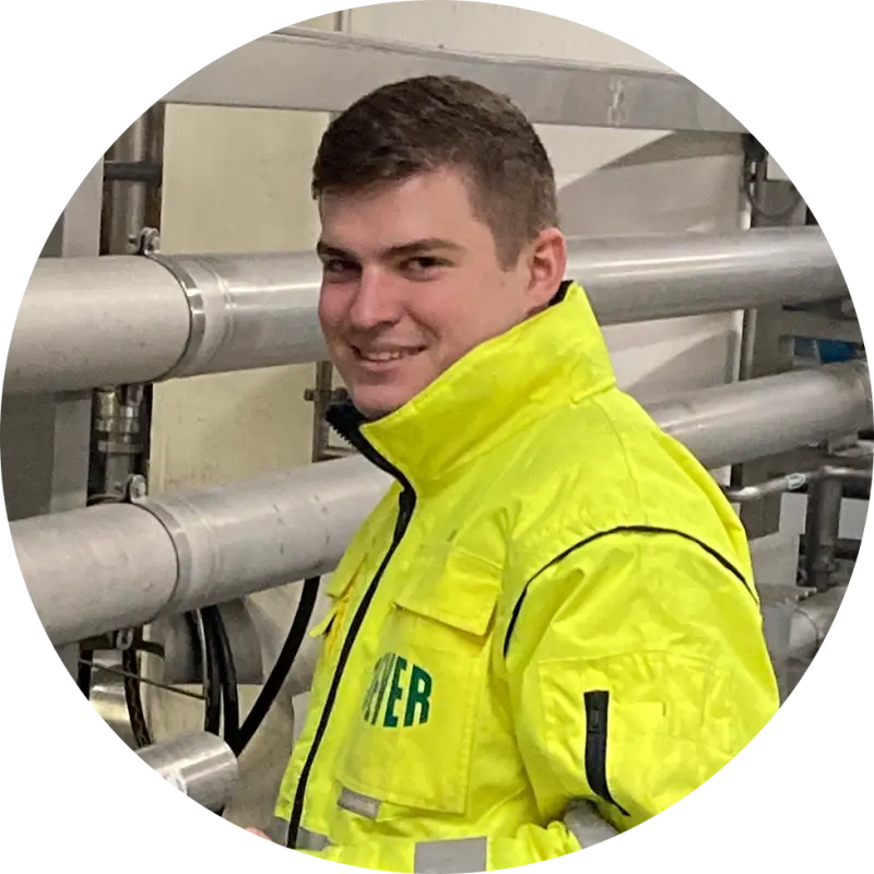 Ein junger Mann in einer neongelben Jacke mit Jever Logo steht in einer Industrieanlage und lächelt.