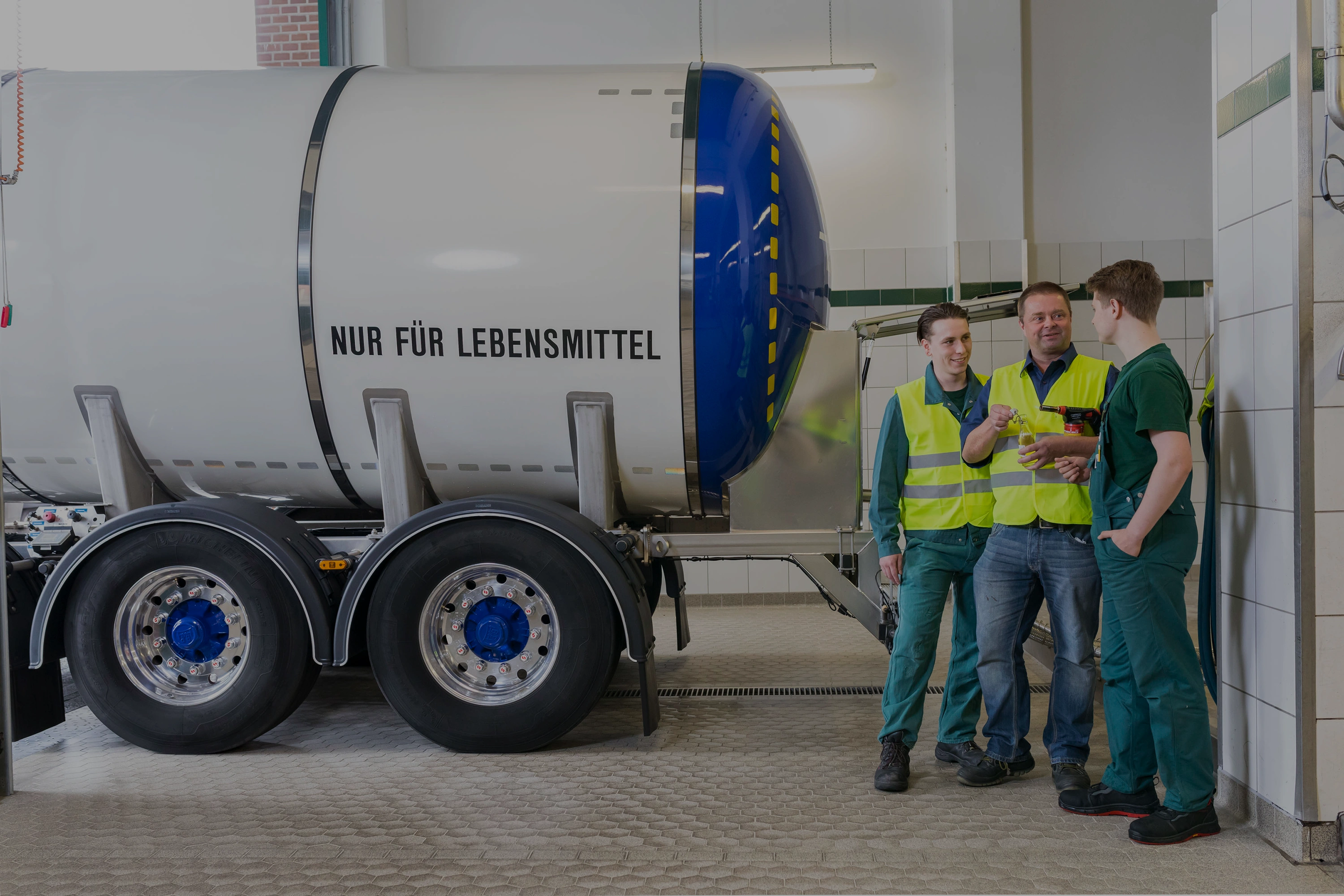 Drei Mitarbeiter sprechen neben einem großen Lebensmitteltank. Zwei tragen Warnwesten, einer hält eine Flasche.