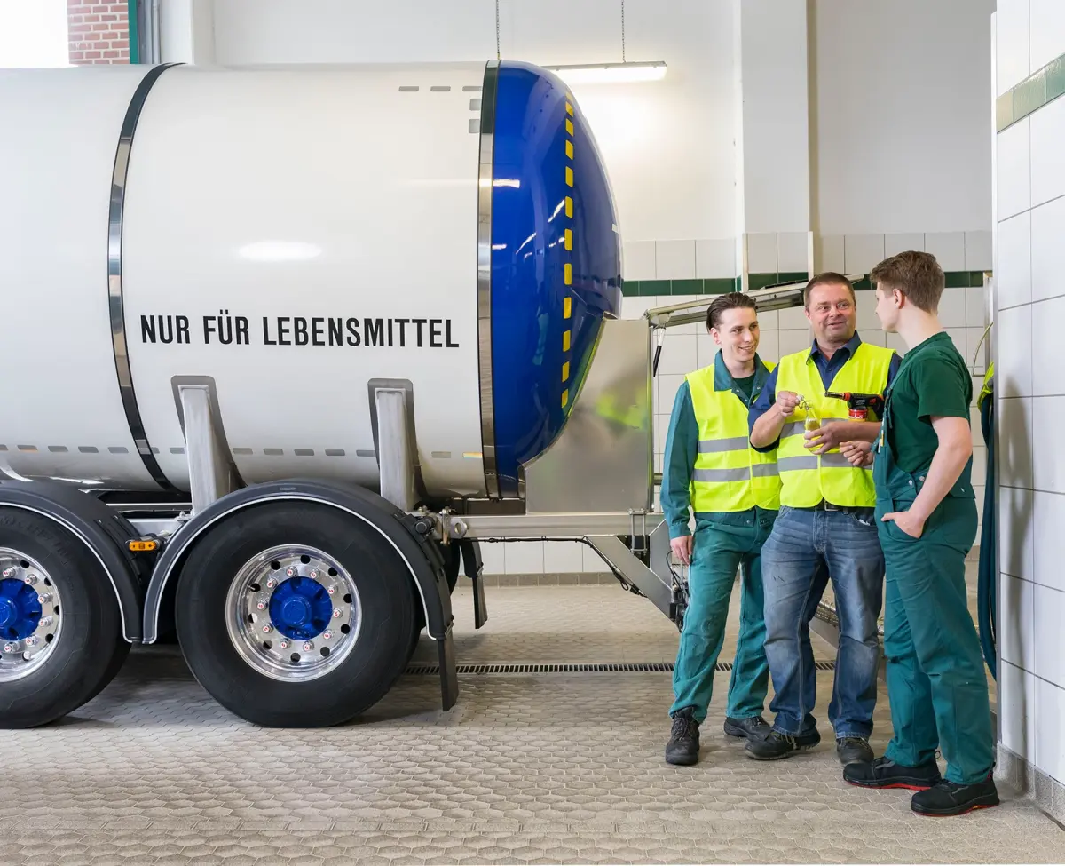 Drei Mitarbeiter sprechen neben einem großen Lebensmitteltank. Zwei tragen Warnwesten, einer hält eine Flasche.