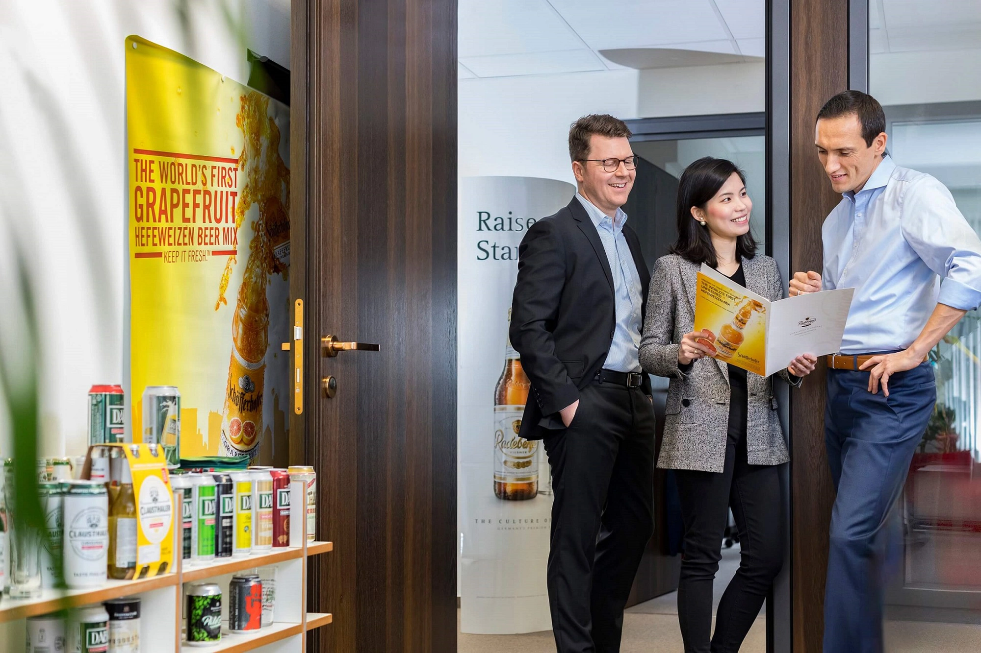 Drei Geschäftsleute stehen lächelnd in einem Büroflur und betrachten Dokumente.