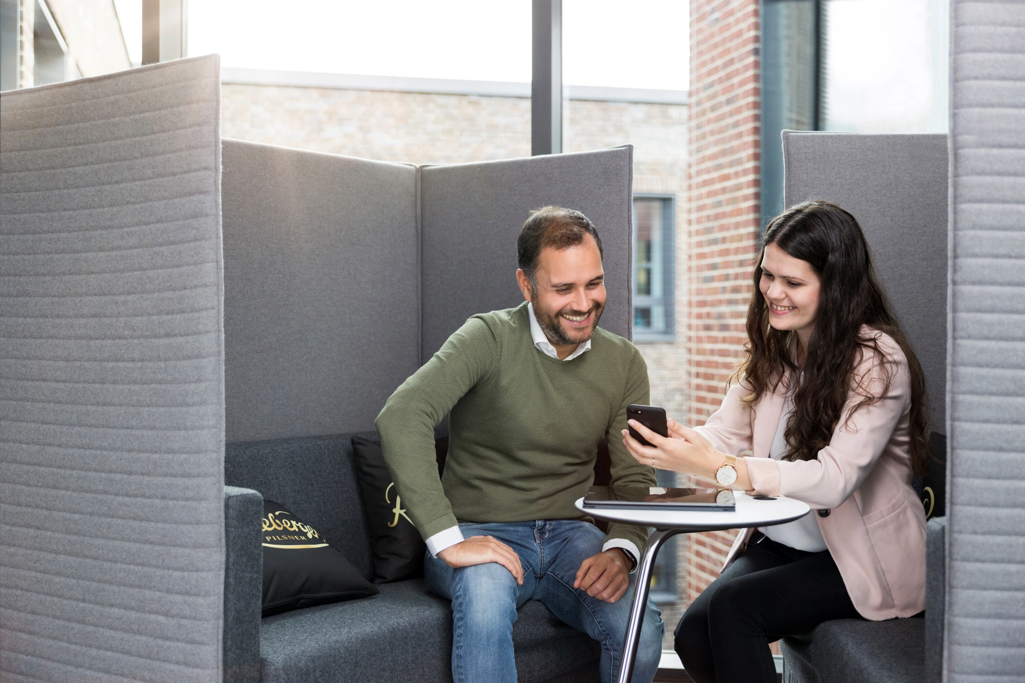 Eine Frau und ein Mann sitzen in einer Büroecke und betrachten lächelnd ein Smartphone.
