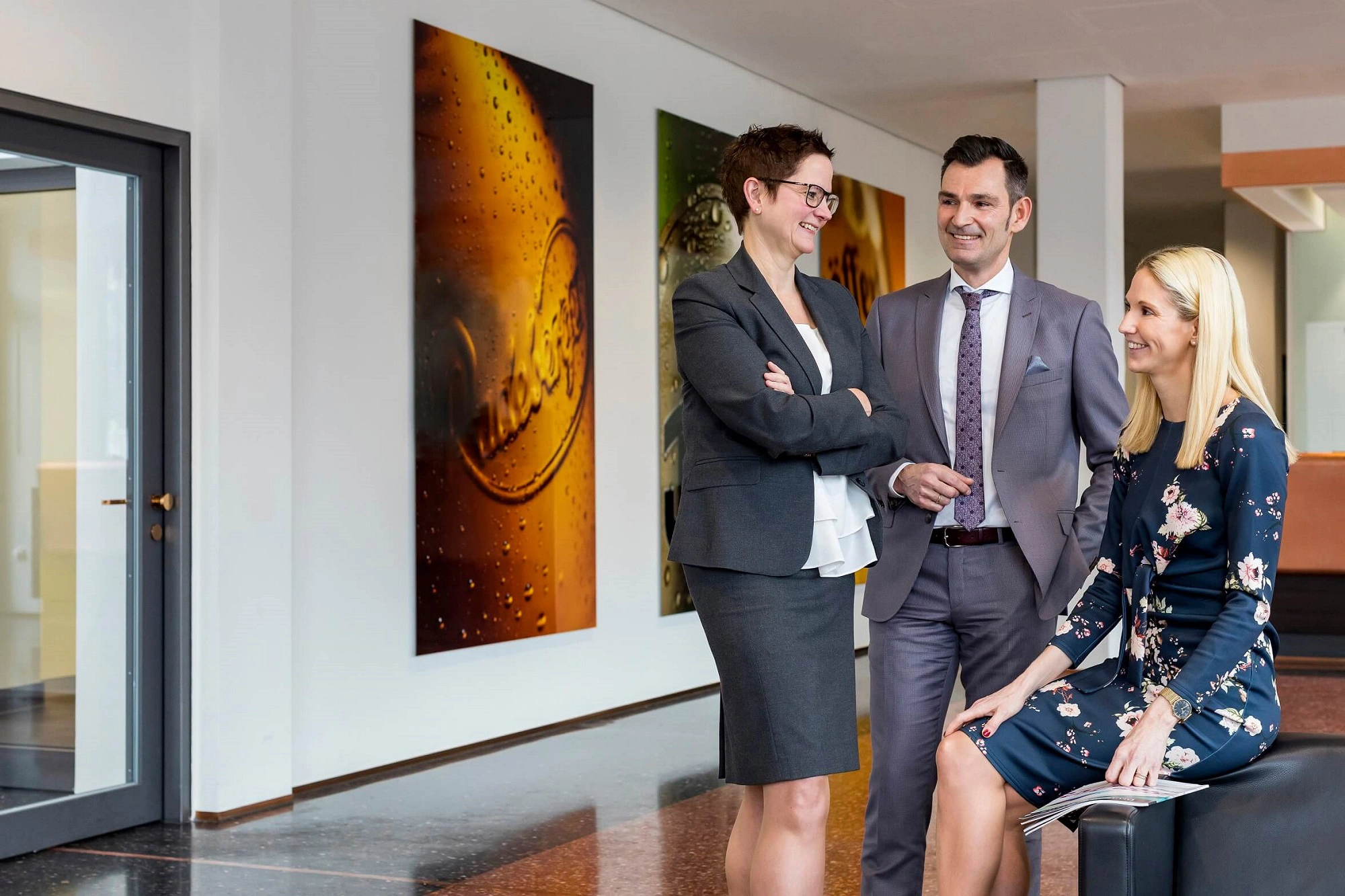 Zwei Frauen und ein Mann stehen lächelnd in einem Büroflur. Die Person rechts sitzt auf einem Hocker.