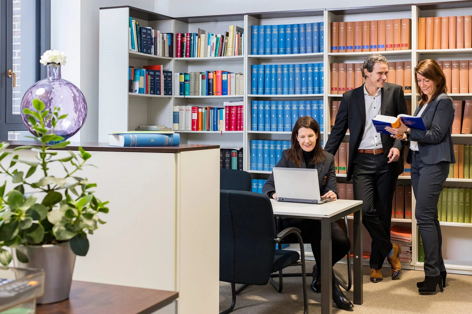 Drei Geschäftsleute besprechen Unterlagen in einem Büro mit vielen Bücherregalen.