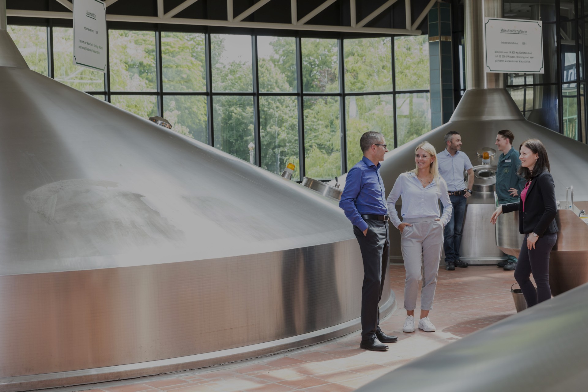 Vier Personen stehen in einer Brauerei und unterhalten sich neben großen Metallkesseln.