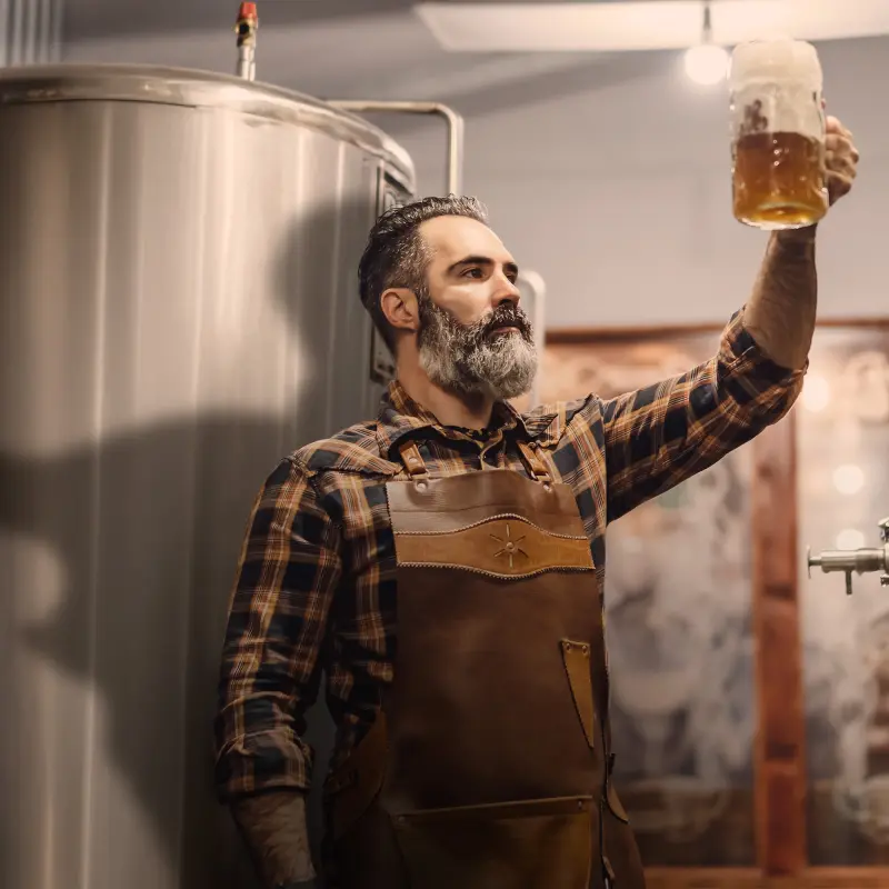 Brauer inspiziert ein gefülltes Glas Bier vor einem großen Edelstahltank in der Brauerei.